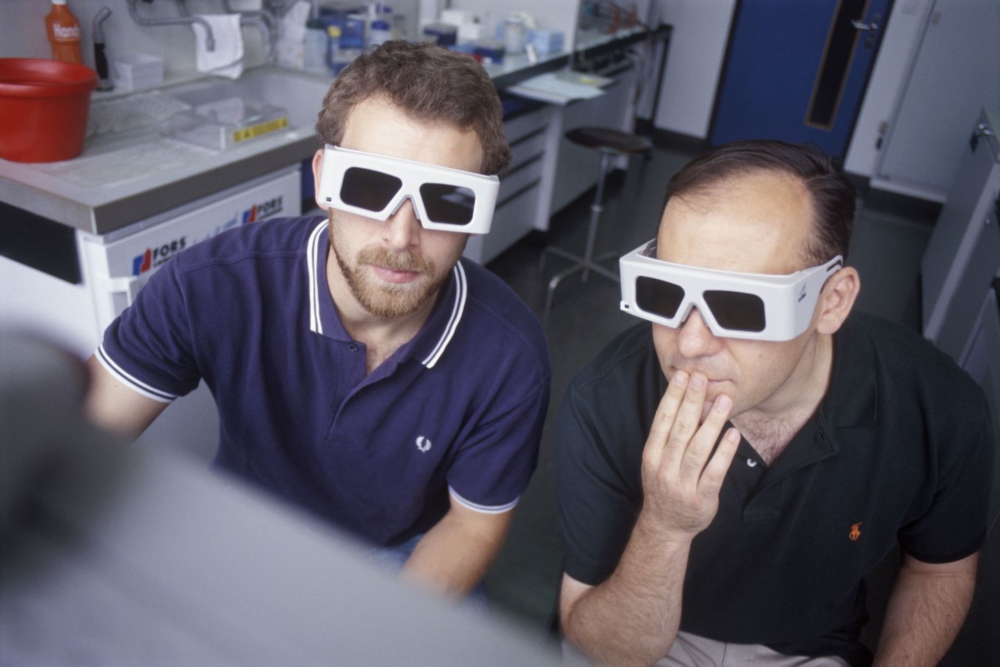 Zwei Männer sitzen in einem Forschungslabor und tragen weisse Brillen. Sie blicken gemeinsam auf einen Computer. 